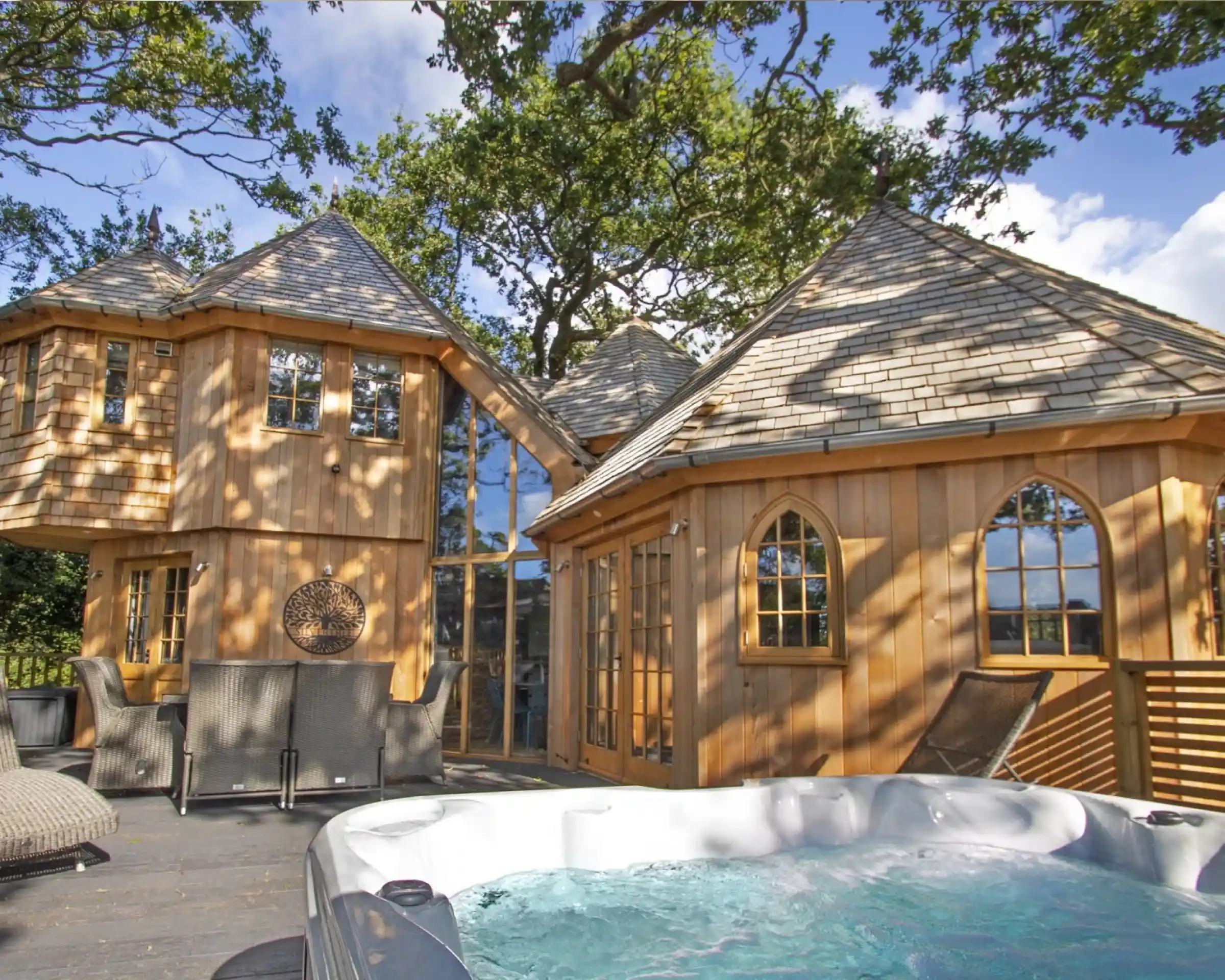 A wooden cabin with unique architecture featuring multiple pointed roofs and large glass windows. A hot tub is positioned on a deck in the foreground, surrounded by lush trees and a blue sky.