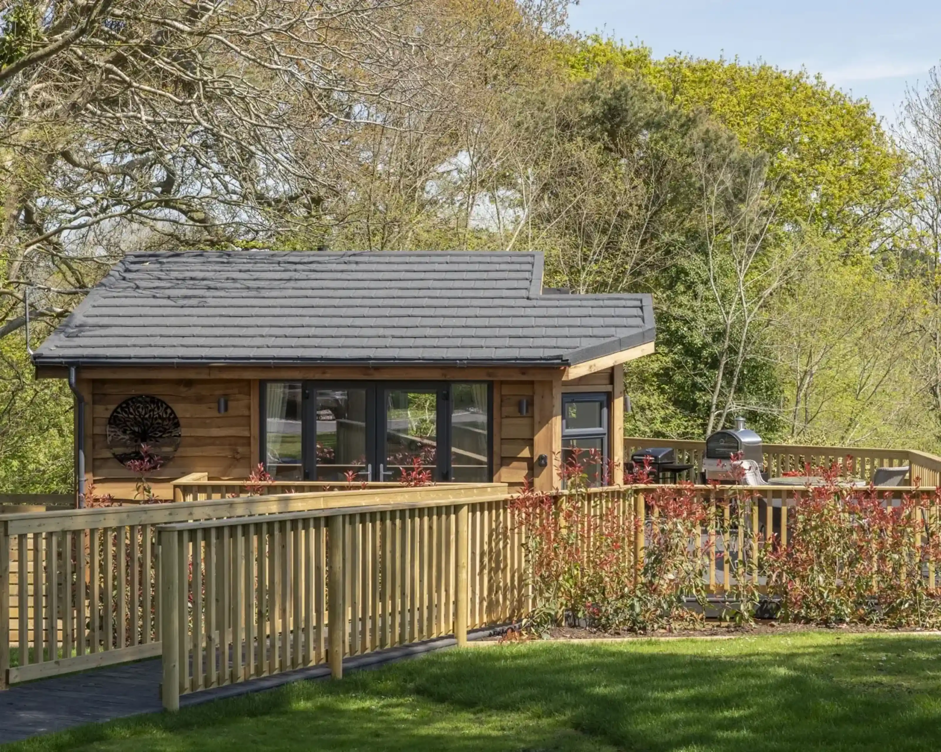 A modern wooden cabin with a gray roof is set against a backdrop of lush greenery. The cabin features large windows and a wooden deck with a railing. A flowering bush is visible in front, enhancing the serene landscape.