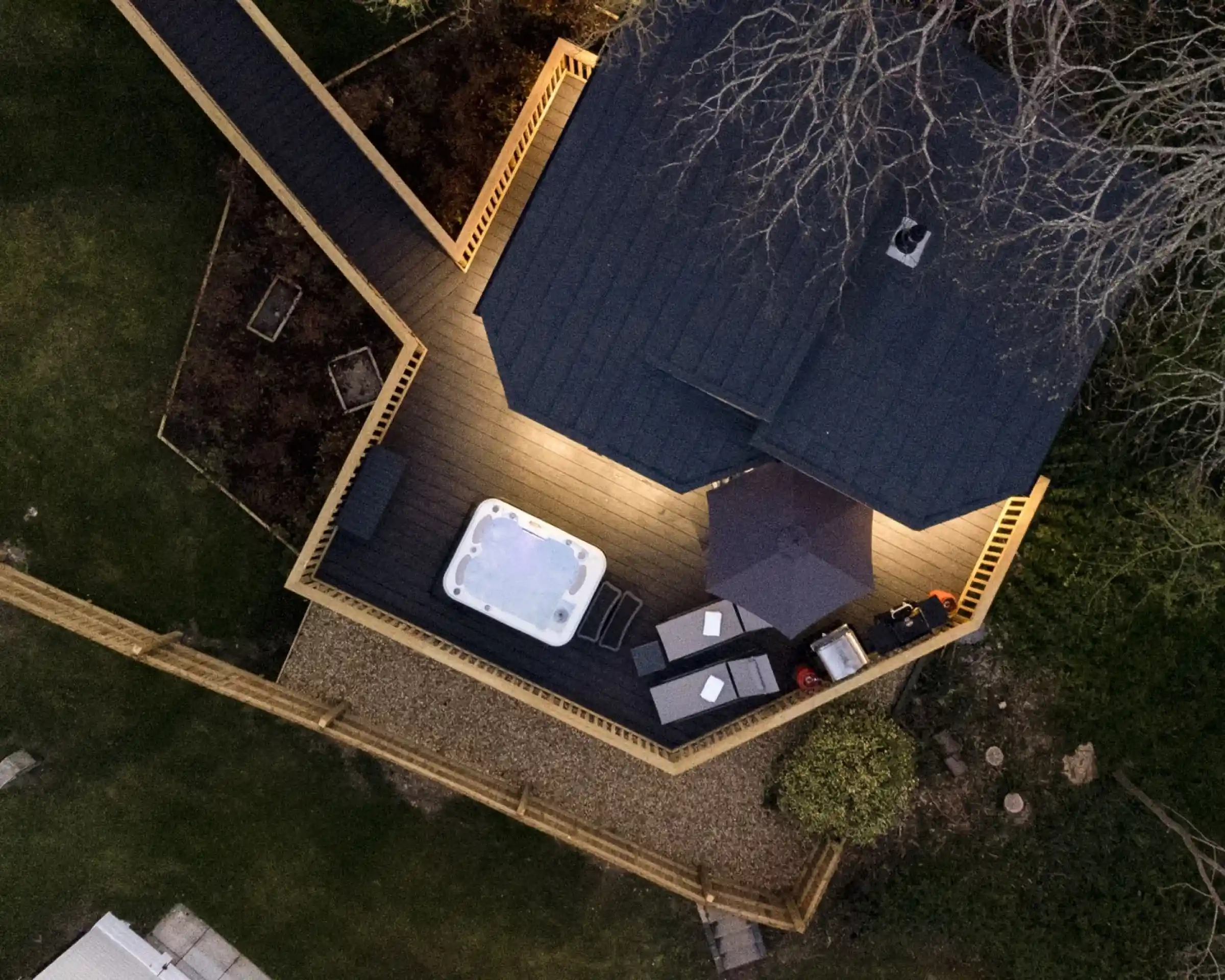 An aerial view of a backyard with a wooden deck featuring a hot tub, outdoor seating area, and an umbrella. The space is illuminated by ambient lighting, surrounded by a well-maintained lawn and garden beds.