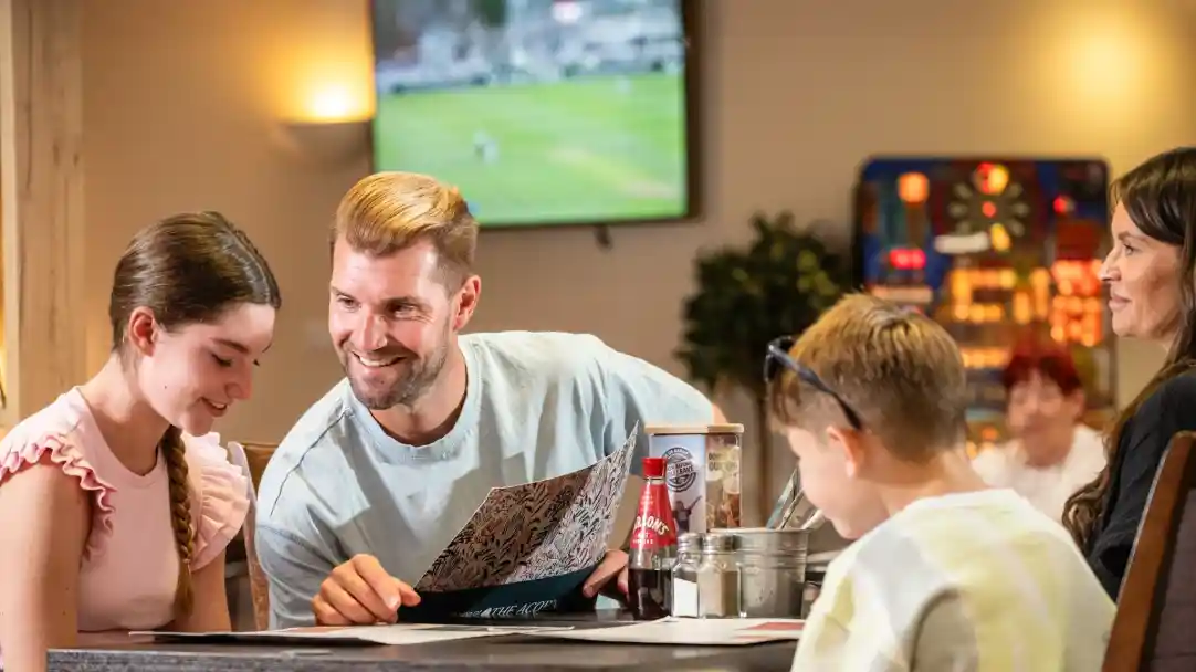 A man and a girl sit at a table, looking at a menu, while a woman and a boy watch. The setting is a casual dining area with a television showing a sports game in the background.