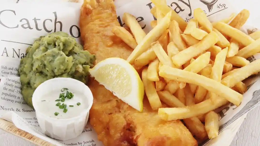 A serving of fish and chips, featuring a golden-battered fish fillet, crispy fries, a side of mushy peas, and a lemon wedge. A small cup of tartar sauce garnished with chives is also included.