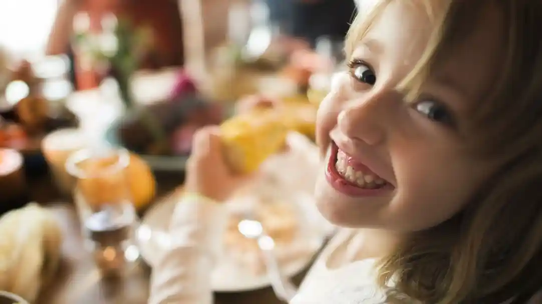A young girl with blonde hair smiles brightly while holding a piece of corn. She is seated at a table filled with food, enjoying a cheerful moment.