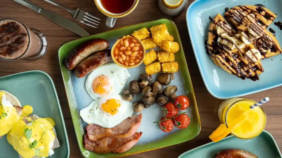 A colourful breakfast spread featuring a plate with two fried eggs, bacon, sausages, baked beans, sautéed mushrooms, and cherry tomatoes. Nearby, a serving of golden hash browns and a plate of waffles drizzled with chocolate sauce. Beverages include coffee and a tropical drink with a straw.