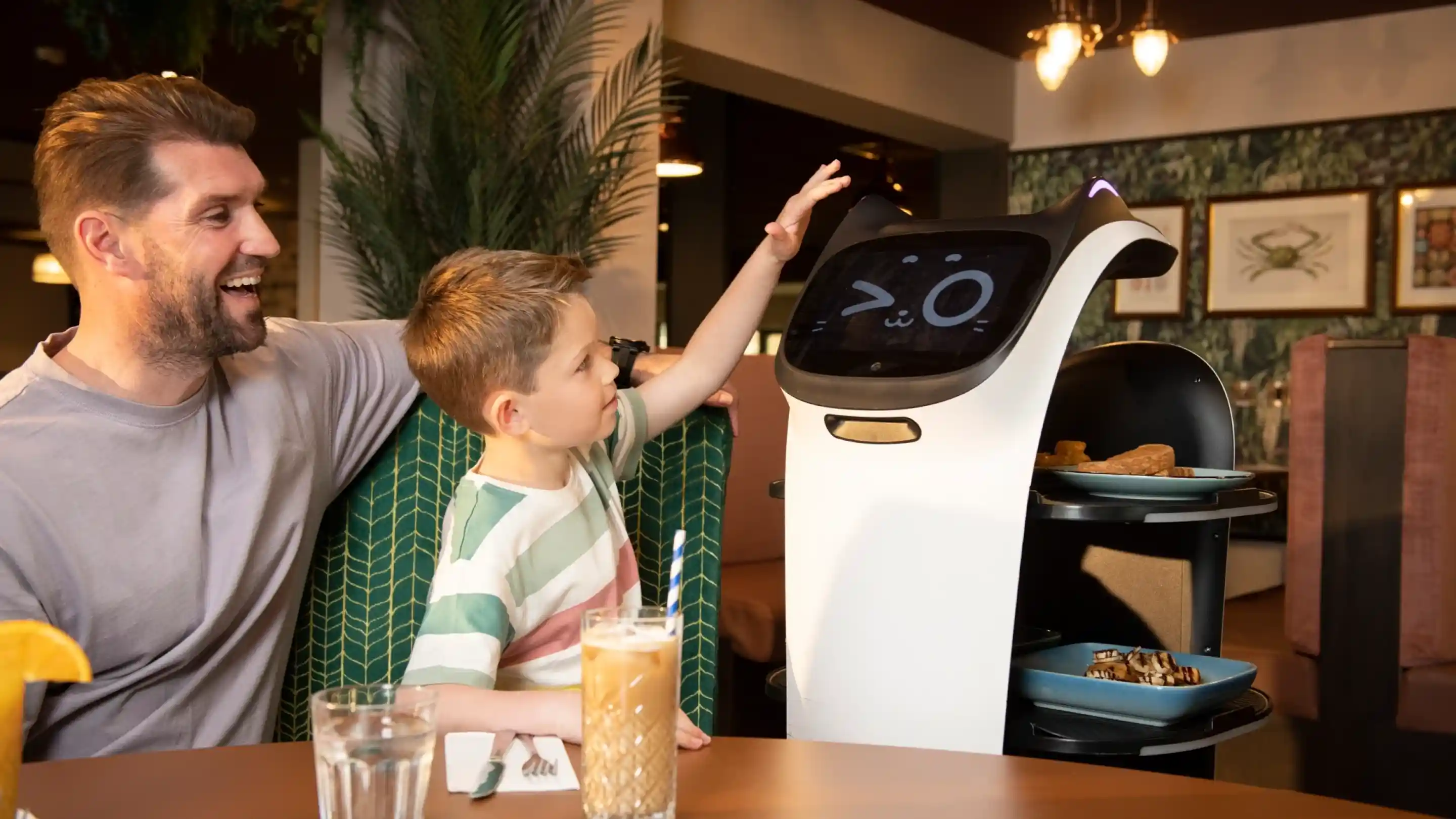 A father and son interact with a friendly-looking, humanoid robot in a restaurant. The child is reaching out to touch the robot's screen, which displays a cute face, while the father smiles beside him. The table is set with food and drinks, and the background features tropical décor.