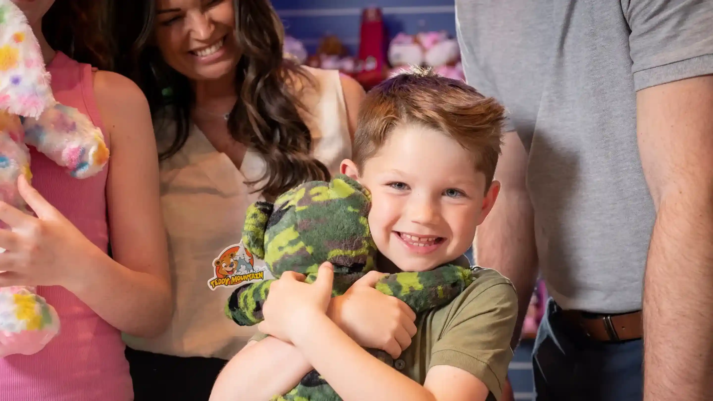 A young boy smiles brightly while hugging a stuffed animal, wearing a camouflage pattern. He is surrounded by other people, including an adult woman visibly delighted. The setting appears to be a store filled with plush toys.