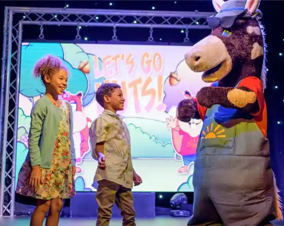 A costumed performer stands on stage with two young children who are smiling and laughing with the performer, with a screen in the background with the words 'Let's Go Nuts!'