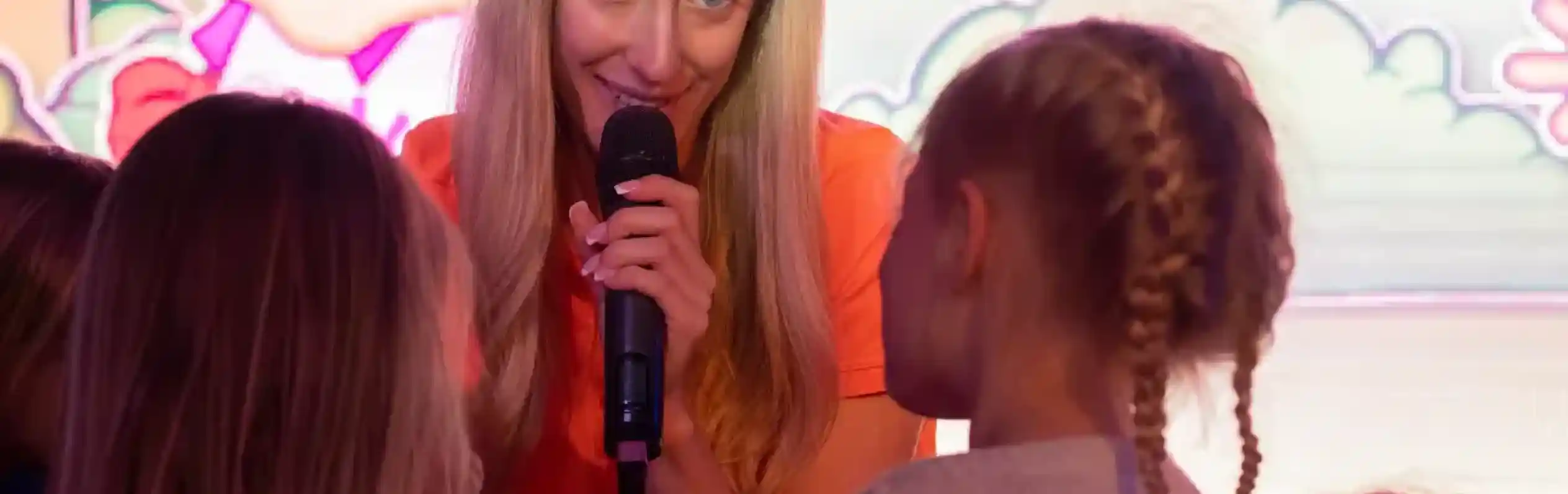 A woman with long blonde hair speaks into a microphone, engaging with a group of young children during an event. A colourful cartoon backdrop features a rabbit character.