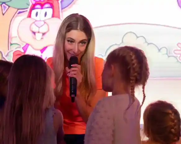 A woman with long blonde hair speaks into a microphone, engaging with a group of young children during an event. A colourful cartoon backdrop features a rabbit character.