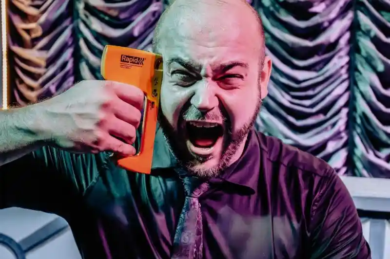 A frustrated man in a black shirt and tie holds an orange stapler to his head, yelling in a dramatic expression against a theatrical backdrop.
