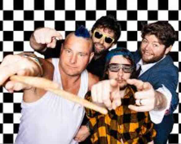 Four men pose in front of a black and white checkered background. One man displays a mohawk hairstyle and is wearing a tank top. Another wears sunglasses and a plaid shirt, while a third sports a blue beanie and sunglasses. The fourth man is dressed in a suit and tie, all are making playful gestures toward the camera.