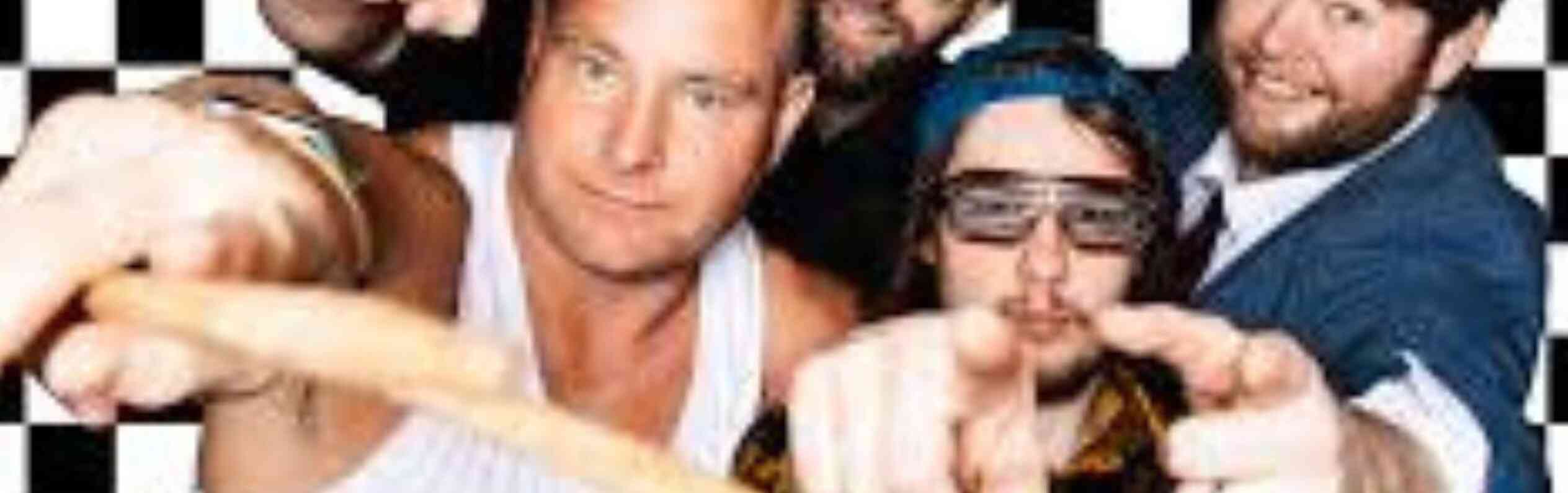 Four men pose in front of a black and white checkered background. One man displays a mohawk hairstyle and is wearing a tank top. Another wears sunglasses and a plaid shirt, while a third sports a blue beanie and sunglasses. The fourth man is dressed in a suit and tie, all are making playful gestures toward the camera.