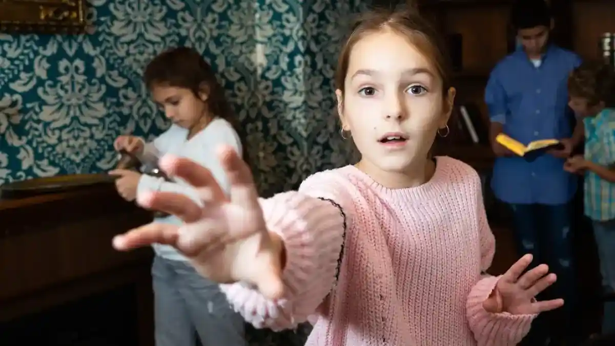 A young girl in a pink sweater reaches out with one hand, looking surprised or curious. In the background, two other children are engaged with books, one focusing on reading while the other appears to be using a tool. The setting features a patterned blue wall and wooden furnishings.