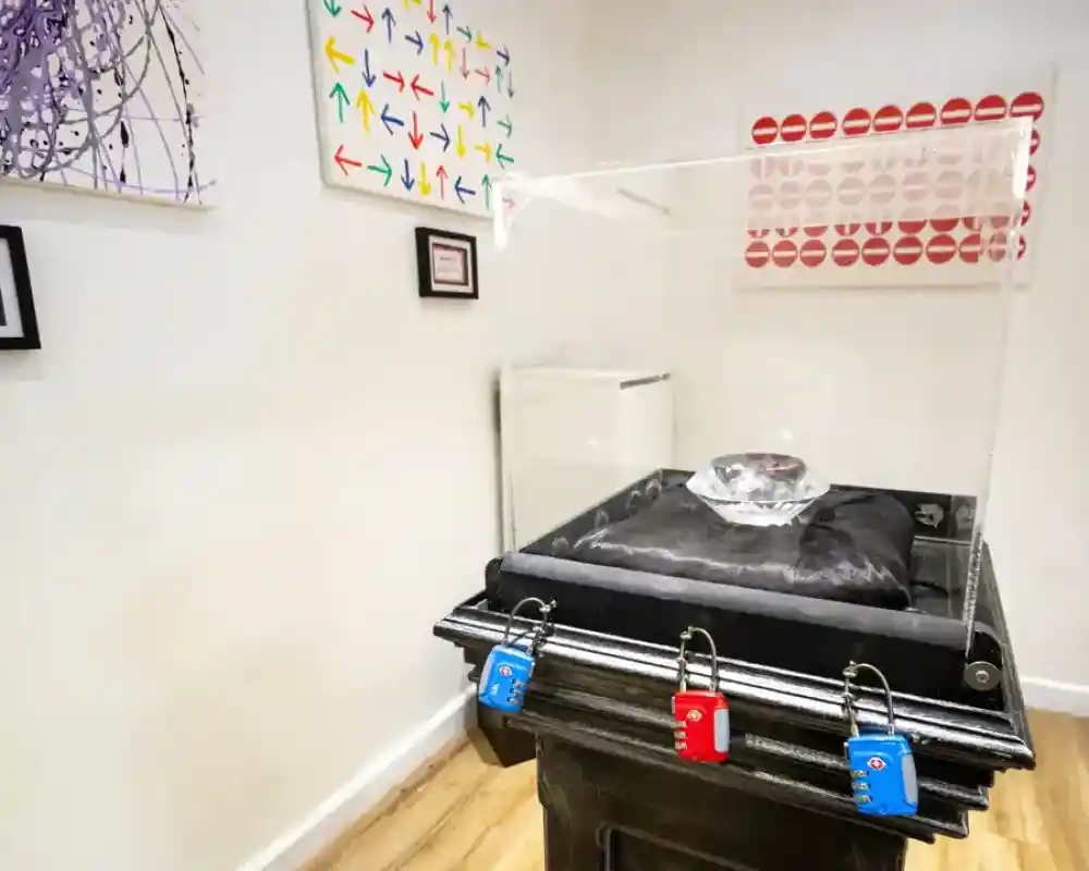 A clear display case surrounds a large, faceted crystal on a velvet cushion, positioned atop a black pedestal. Nearby walls feature colourful abstract art, including patterns of arrows and lip prints, framed in black. Three locks in blue and red secure the display.