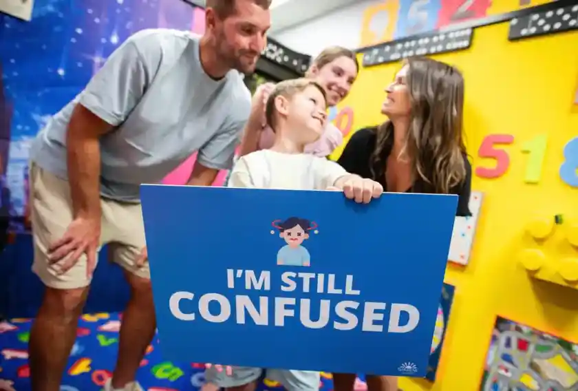 A group of four people stands in a colourful room filled with educational toys and decorations. A child holds a blue sign that says, I'm still confused. The adults, smiling and looking at the child, convey a sense of playful engagement.