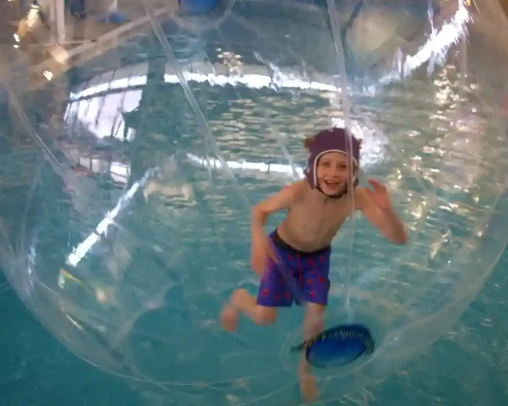 A young boy inside a transparent inflatable bubble, floating on the water surface of a pool. He is smiling and waving, wearing swim shorts and a swim cap.