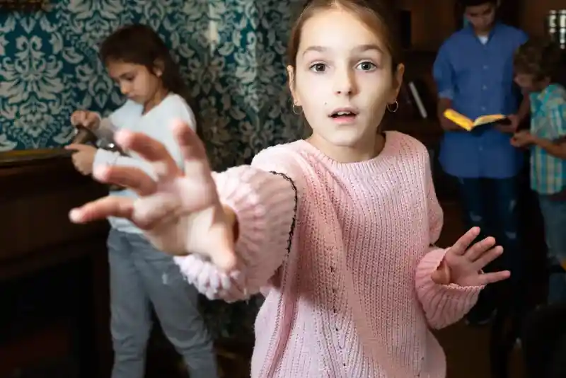 A girl in a light pink sweater reaches out towards the camera with a surprised expression. In the background, two other children are engaged in activities: one is reading a book while the other is focused on a task. The setting has ornate wallpaper and a cosy atmosphere.