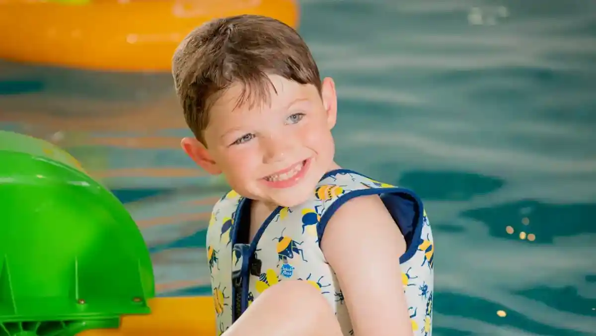 A smiling boy wearing a colourful life jacket sits on a floating platform in a swimming pool. He has light brown hair and appears joyful, with vibrant green inflatable wheels nearby.