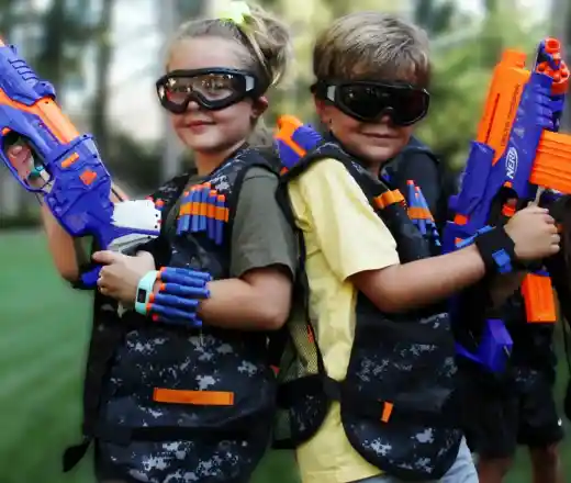 Two children stand back-to-back, holding colourful toy blasters. They wear tactical vests and protective goggles, with a bright outdoor setting in the background.