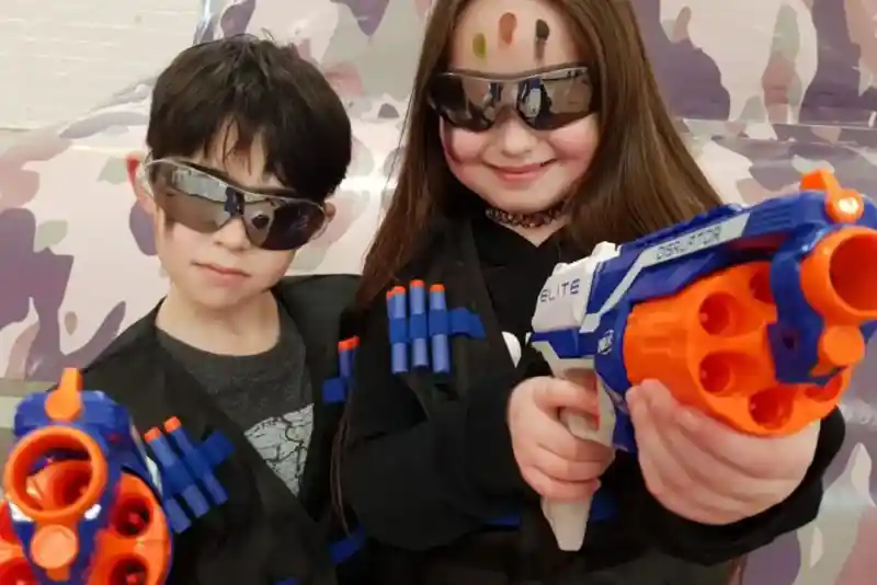 Two children dressed in tactical vests with toy guns. They wear sunglasses and have colourful face paint on their foreheads. The background features a camouflage design.