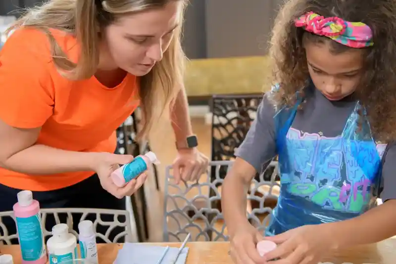 A woman in an orange shirt guides a young girl with curly hair and a colourful headband as they mix paint at a table. The girl is focused on her work, while the woman holds a bottle of paint, providing instructions. Various art supplies are spread out on the table.