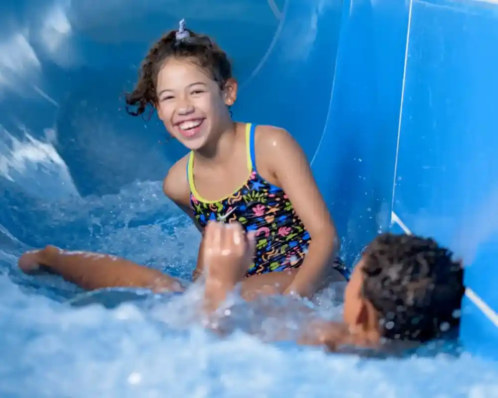 A young girl in a colourful swimsuit laughs as she rides down a water slide, while a boy splashes in the water below her. Both are enjoying a fun day at a water park.