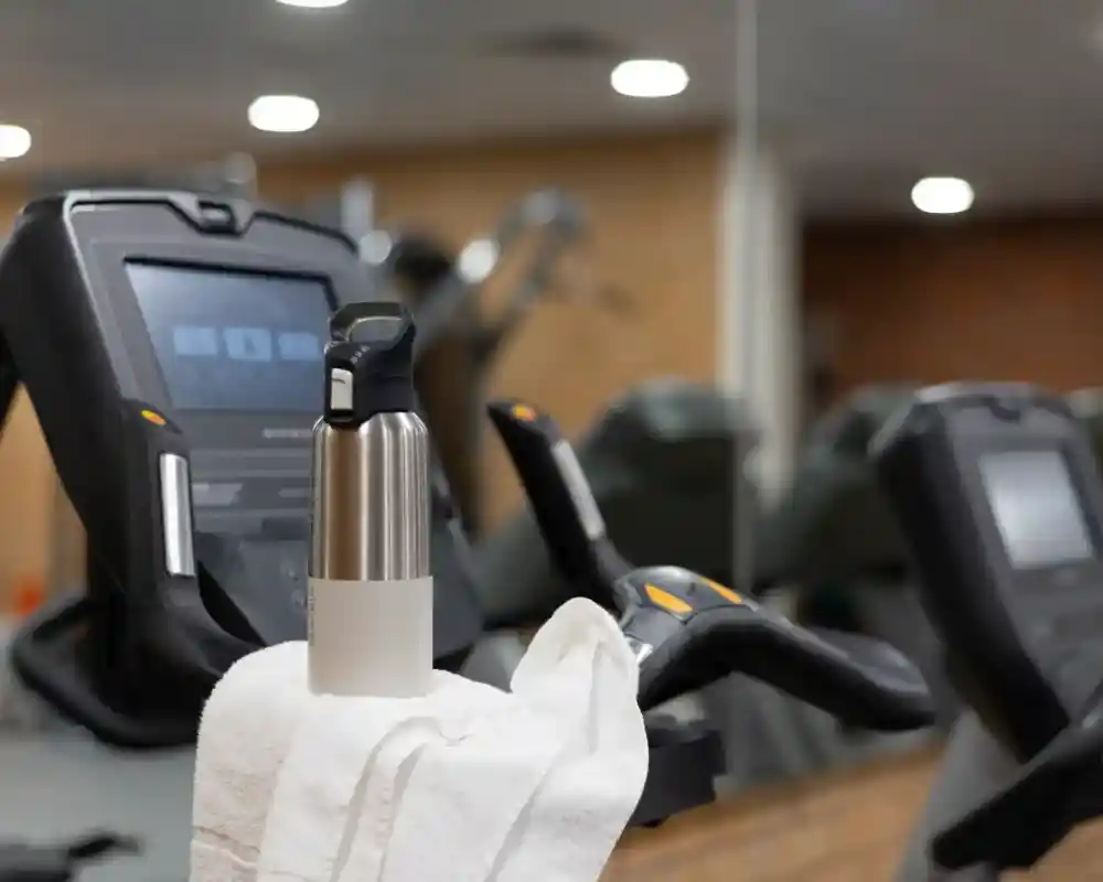 A stainless steel water bottle sits atop a white towel on the handlebars of an exercise machine in a gym, with blurred workout equipment in the background.