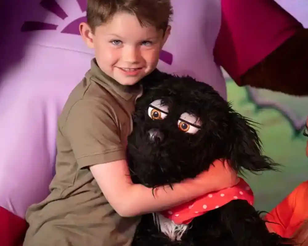 A young boy smiles while hugging a fluffy black puppet with large eyes and a red scarf. In the background, a cartoon character in a pink shirt is partially visible.