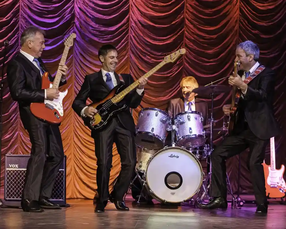 A band of four musicians performing on stage, dressed in suits. Two guitarists play electric guitars while a bassist stands in the center, smiling. A drummer sits behind a drum kit, contributing to the lively atmosphere with colorful stage lighting in the background.