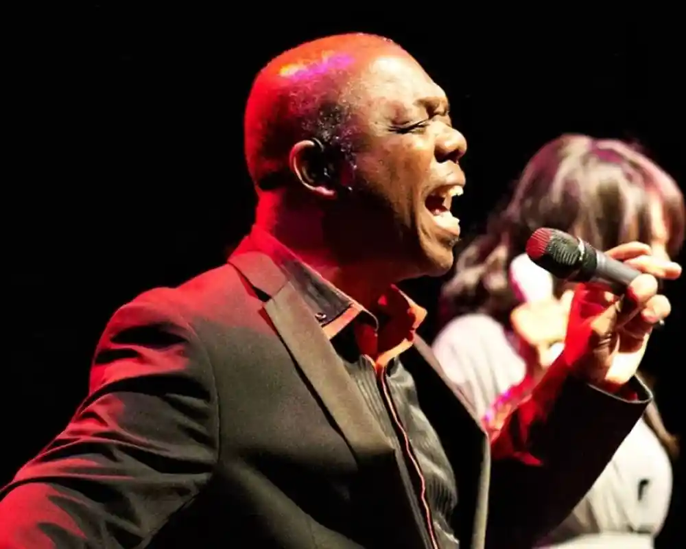 A man passionately singing into a microphone on stage, wearing a black suit with a red shirt. He has a focused expression and is illuminated by stage lighting, while a woman in the background is partially visible.