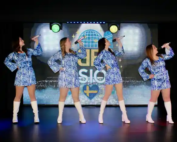 A group of four women performing on stage, each wearing matching blue patterned outfits and white knee-high boots. They hold microphones and pose confidently, with colorful stage lights in the background.