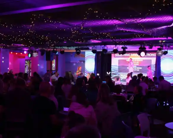 A lively indoor venue filled with people enjoying a performance. The stage is illuminated with colorful lights, and a large screen displays text. String lights hang overhead, creating a festive atmosphere.