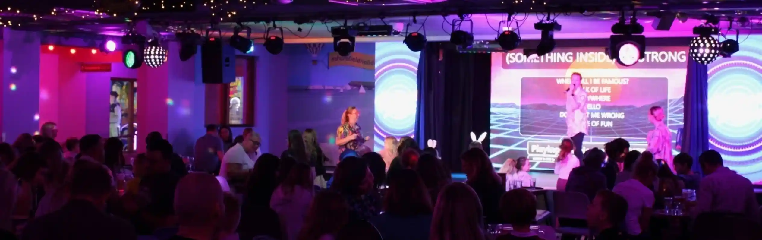 A lively indoor venue filled with people enjoying a performance. The stage is illuminated with colorful lights, and a large screen displays text. String lights hang overhead, creating a festive atmosphere.