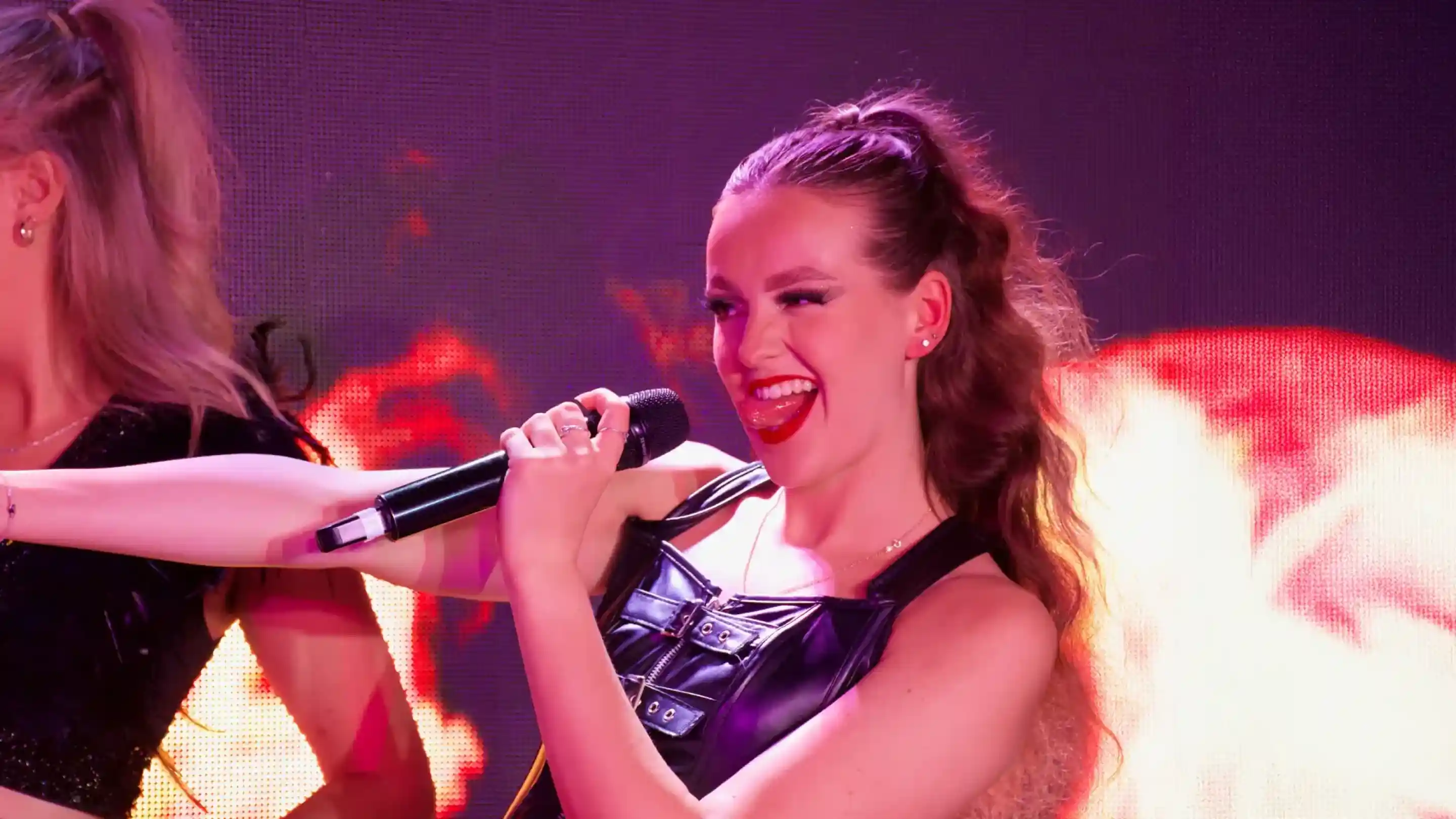 A performer with wavy hair holds a microphone while singing, wearing a black leather outfit. She has a playful expression and bright red lipstick, with a fiery background enhancing the energetic atmosphere.