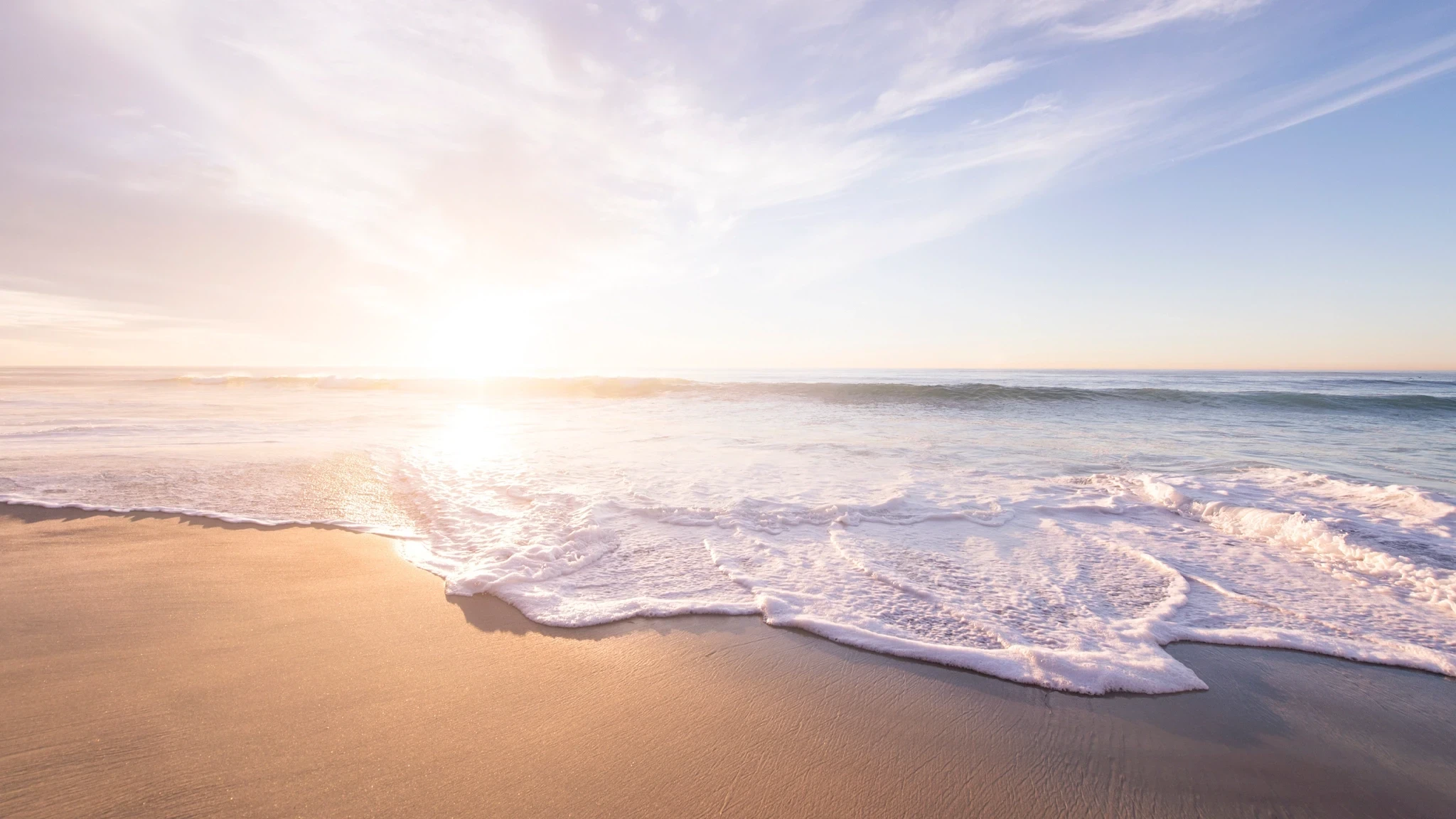Gentle waves wash onto a sandy beach at sunset, with soft pastel colours in the sky and sunlight reflecting on the water.