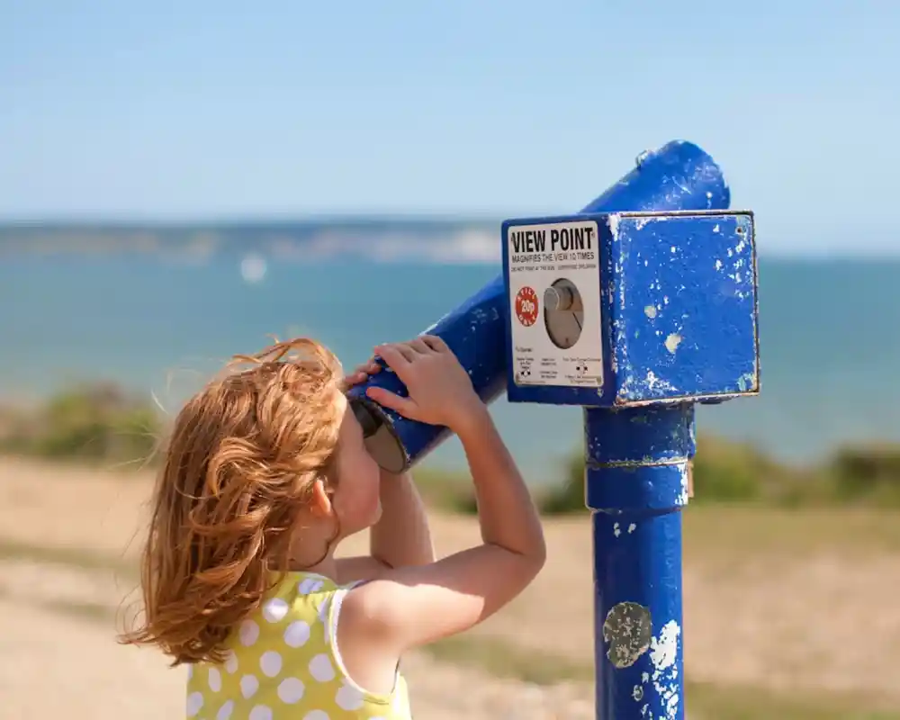 A young girl with red hair wearing a yellow and white polka dot dress is looking through a blue viewpoint telescope, with a scenic view of the ocean in the background.