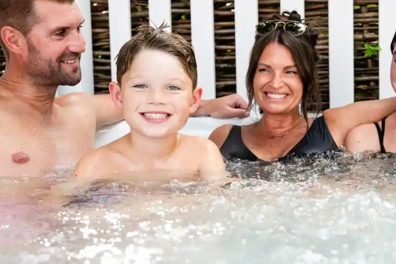 A smiling young boy sits in a hot tub with two adults, one man and one woman, both smiling back at him. The setting has a relaxed, cheerful atmosphere, with a wooden fence in the background.