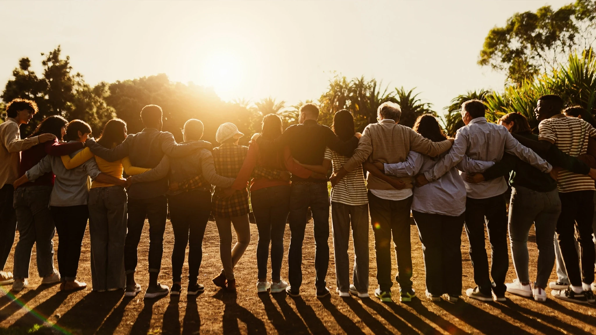 A group of diverse individuals stands together with their arms around each other, facing a sunset. They are positioned in a line, with shadows stretching behind them, against a backdrop of palm trees and a warm, glowing sky.