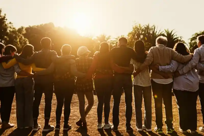 A diverse group of people stands together, linking arms and facing a sunset. They are outdoors, surrounded by trees, with warm sunlight illuminating the scene.