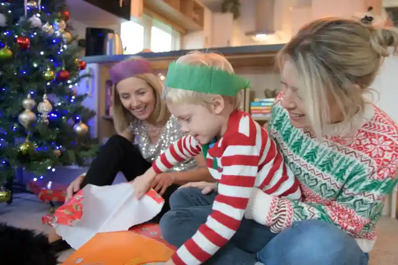 Two women and a young boy are sitting on the floor beside a Christmas tree. The boy, wearing a striped sweater, is excitedly unwrapping a gift, while the women smile and watch him. Festive decorations and a colourful background enhance the holiday atmosphere.