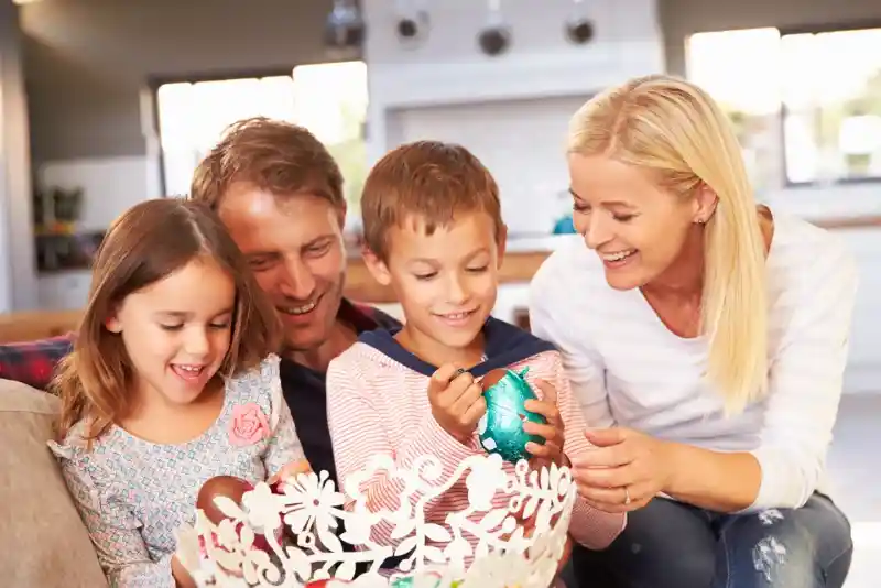A family of four sits together on a couch, joyfully decorating colourful Easter eggs. The parents smile at their two children, a boy and a girl, who are engaged in painting the eggs in a decorative bowl. The bright and inviting living space adds to the cheerful atmosphere.