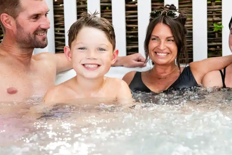 A smiling young boy sits in a hot tub with two women and a man. They are all enjoying the water, with the background showing a wooden fence. The mood is cheerful and relaxed.