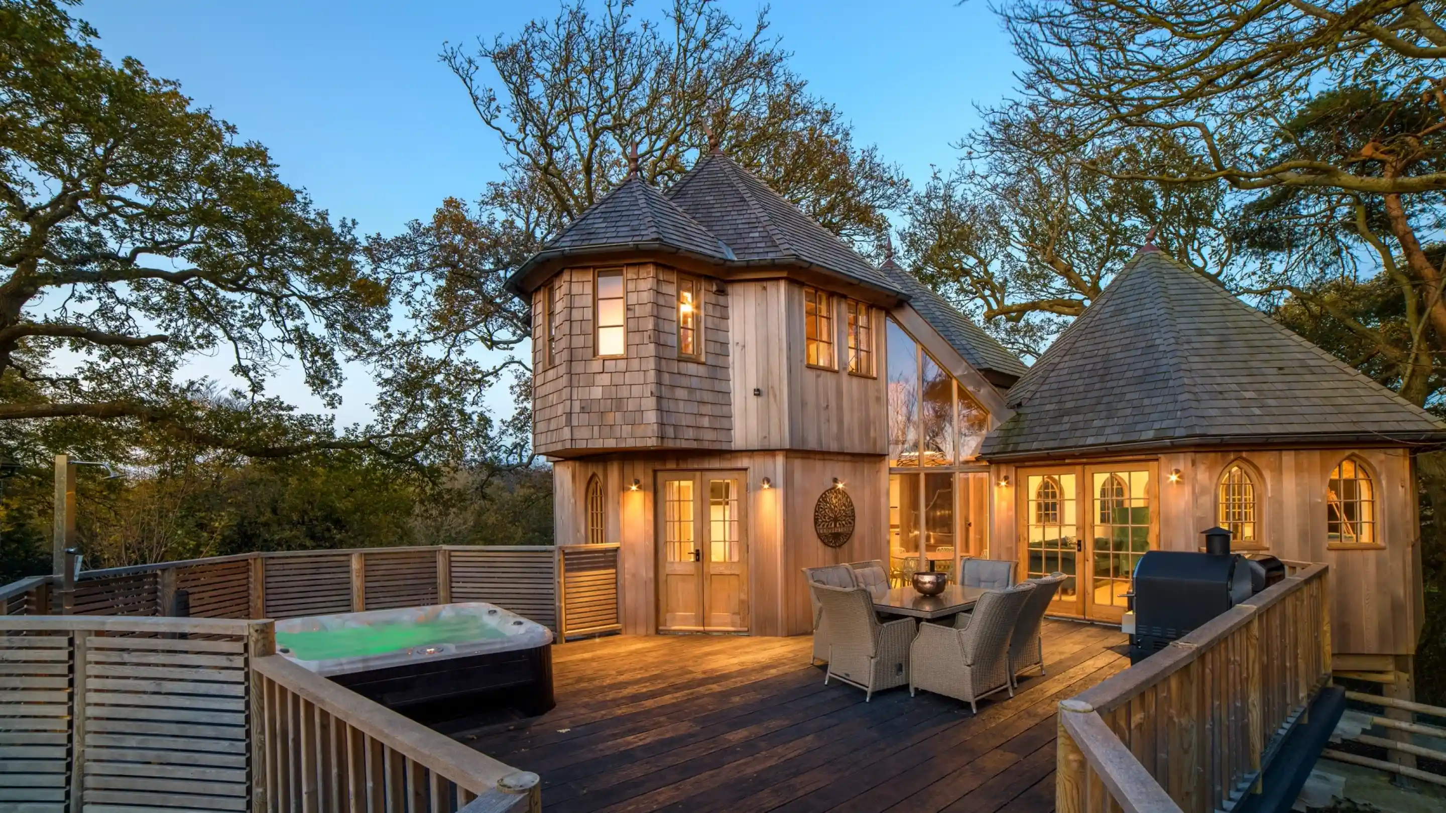 A charming wooden treehouse with multiple towers and a spacious deck, surrounded by trees. A hot tub sits on the deck next to a dining area with wicker furniture. The building features large windows and a cosy design, illuminated by evening light.