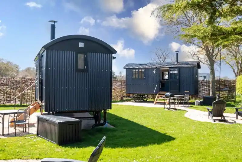 Two stylish shepherd huts situated in a grassy area, surrounded by a wooden fence. One hut is larger with a porch and chimney, while the smaller hut features a curved roof. Adirondack chairs are scattered around for seating. Bright blue sky and fluffy clouds complement the serene outdoor setting.