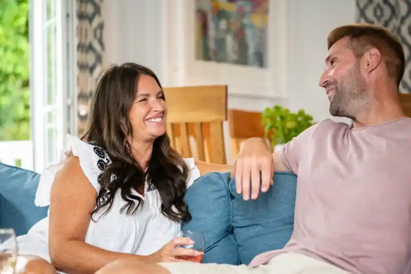 Two people sitting on a blue sofa in a cosy room, engaging in a cheerful conversation. One person has long dark hair and is wearing a white top, while the other has short hair and is dressed in a light pink shirt. A glass is held by the woman, and there are plants and colourful artwork in the background.