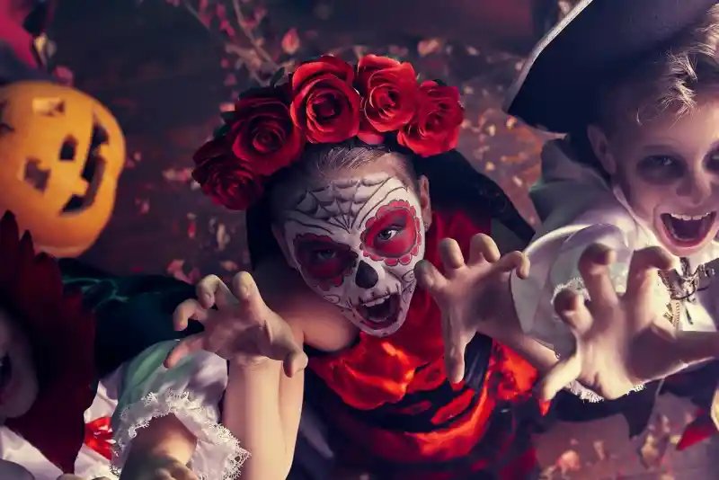 Three kids in costumes pose dramatically for Halloween. One child wears a traditional Día de los Muertos outfit with a skull face paint and flower crown. Another is dressed as a vampire with a cape, while the third is a pirate. They have playful expressions and are reaching toward the camera. Behind them, a pumpkin adds to the festive atmosphere.