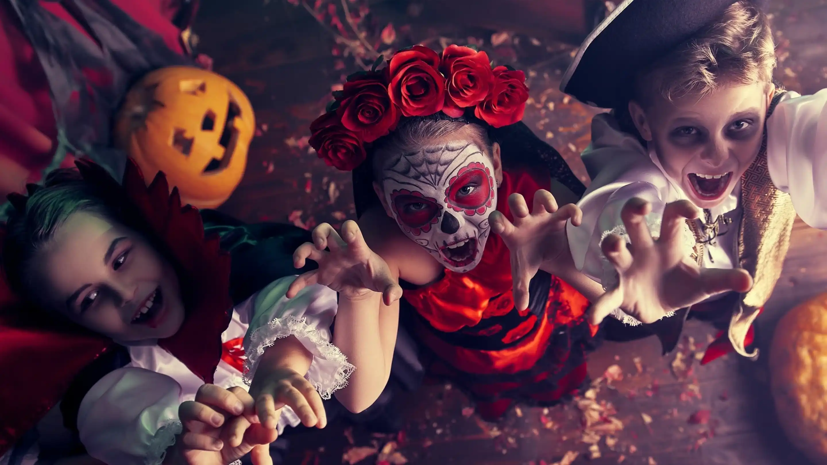 Three children in Halloween costumes reach out playfully towards the camera. One child wears a vampire outfit, another is dressed as a skeleton girl with face paint and a red dress, and the third is in a pirate costume. A carved pumpkin is visible in the background, surrounded by colorful autumn leaves.