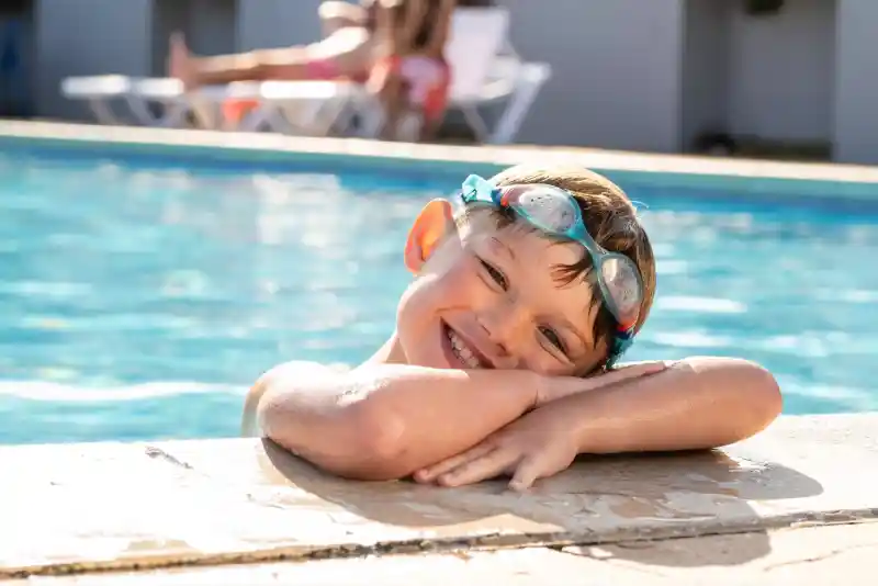 A smiling boy with goggles resting on his head leans on the edge of a swimming pool. In the background, a girl relaxes on a lounge chair by the water. Bright sunlight illuminates the scene.
