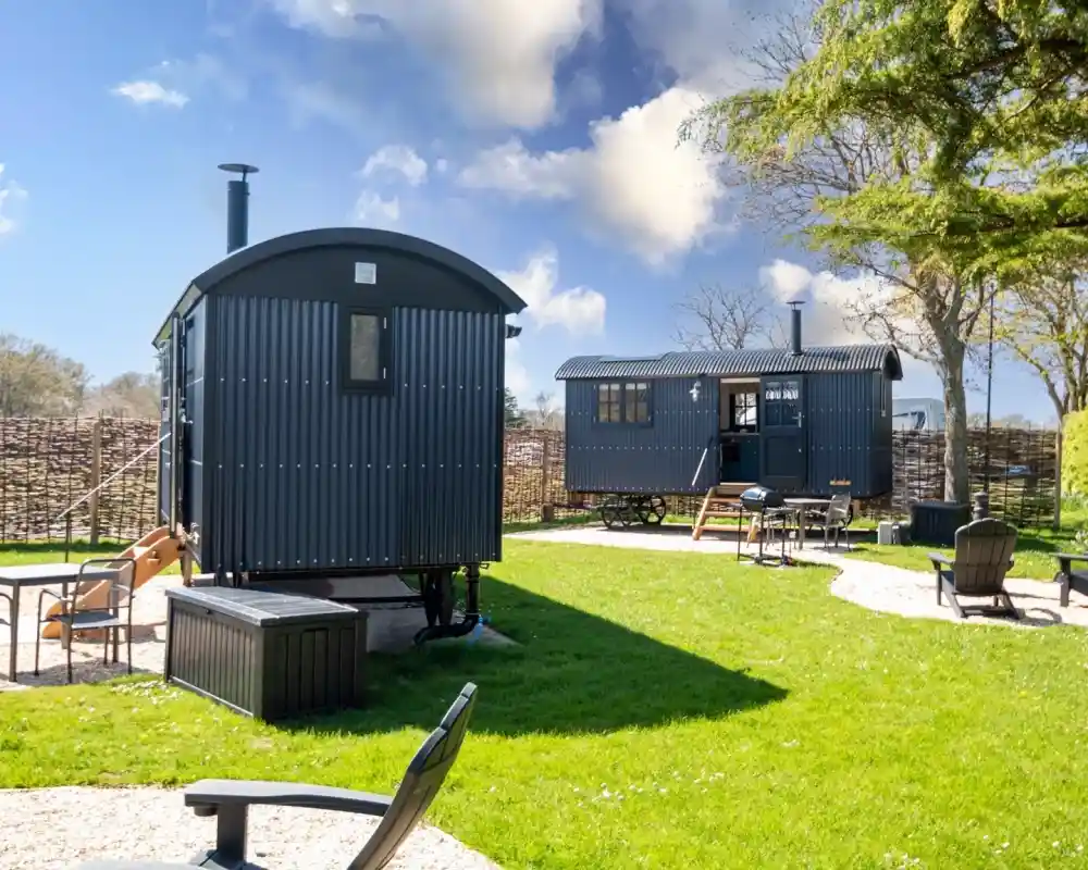 Two stylish shepherd's huts are set in a lush green garden. One hut features a rounded top with a chimney, while the other has a more rectangular shape and a porch. Surrounding the huts are seating areas with patio chairs and a backdrop of trees and a woven fence. The sky is bright with fluffy clouds.