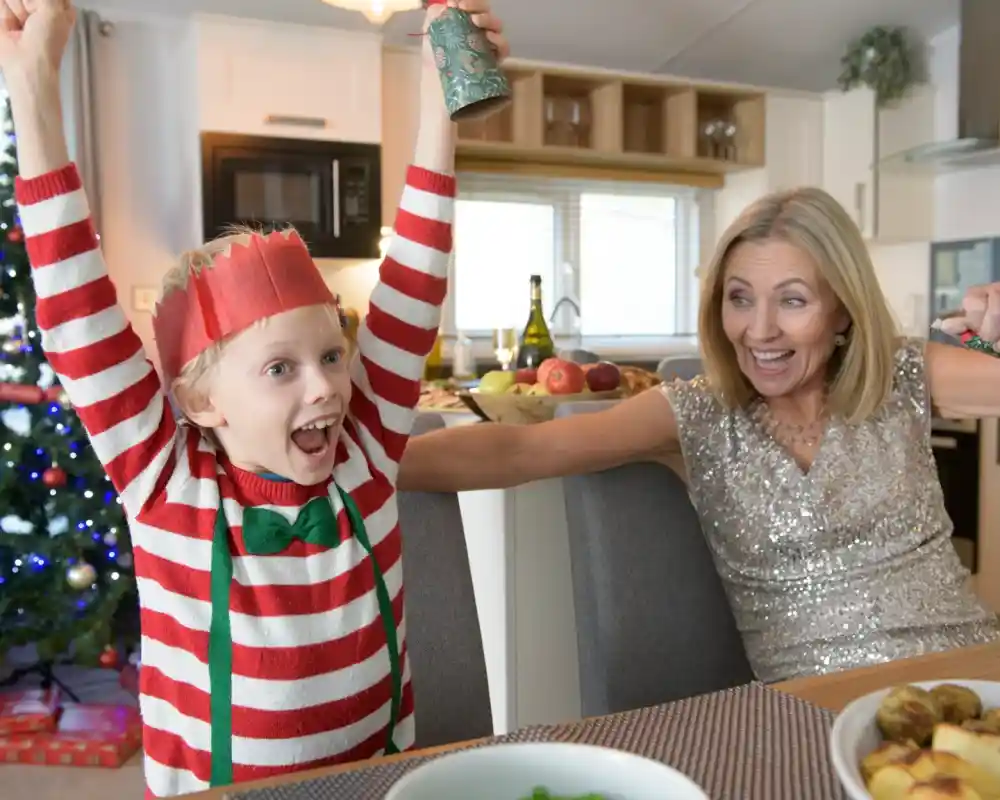 A joyful child in a red and white striped sweater, wearing a paper crown, celebrates with raised arms. A smiling woman in a sparkling dress sits nearby, sharing in the festive excitement, with a decorated Christmas tree visible in the background.