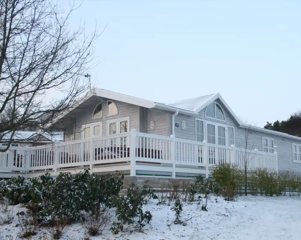 A modern wooden cabin with a large deck, surrounded by snow and bare trees. The cabin features several windows and a gabled roof.